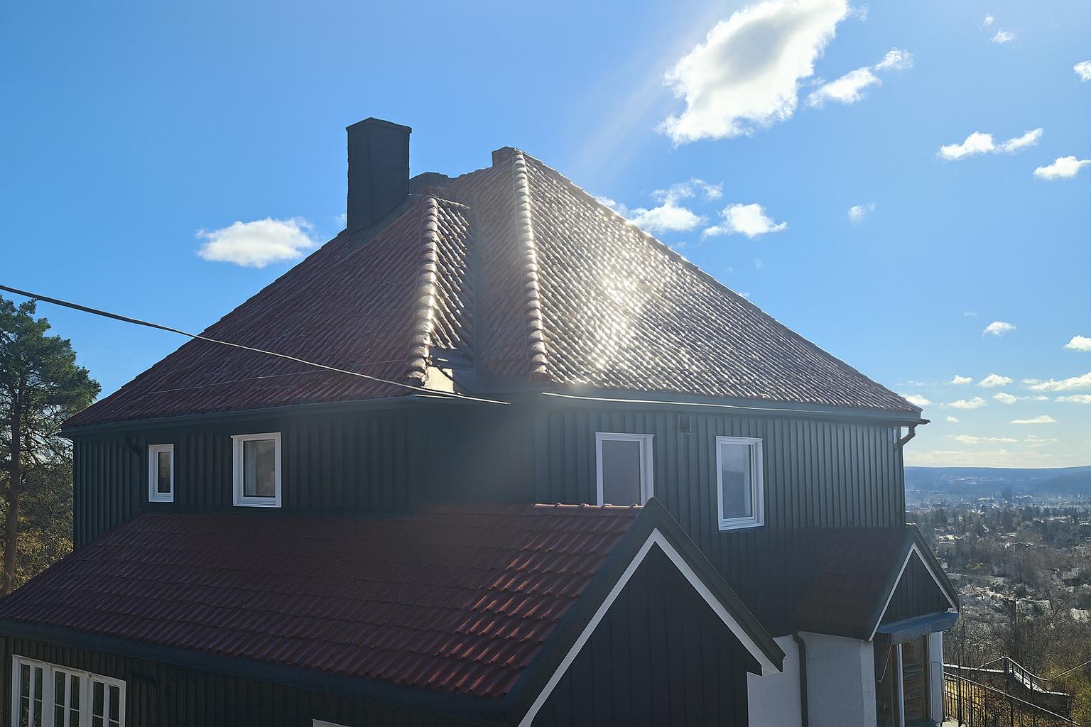 Enebolig med mørk fasade og rødt tak, fotografert i sollys med utsikt over bebyggelse.