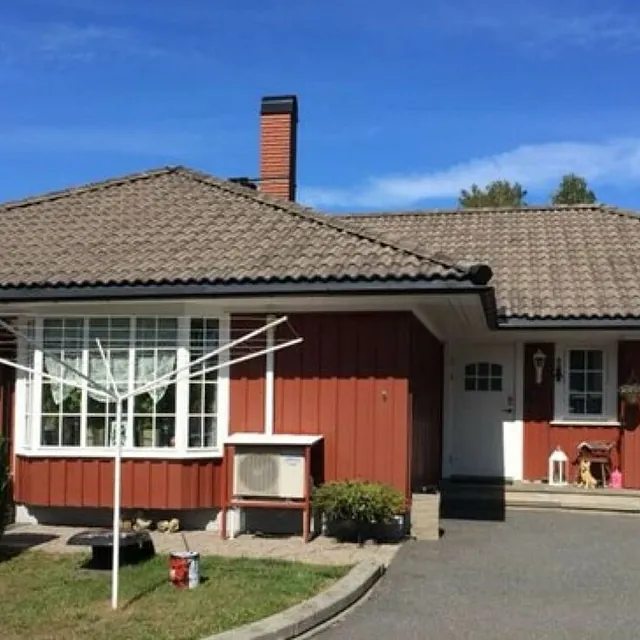 Enebolig med rød trekledning, valmet tak og inngangsparti fotografert i solfylt boligområde.