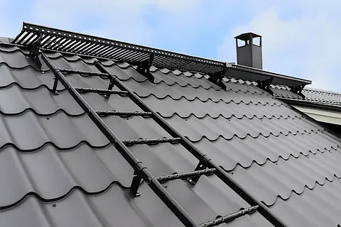 Tak med snøfangere, takstige og pipe montert på grått metalltak med taksteinsprofil.