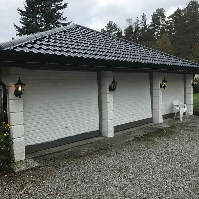 Frittstående garasje med hvit fasade, mørkt metalltak og utelamper i gruslagt gårdsrom.