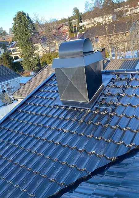 Takhatt i sort metall montert på mørkt tak med taksteinprofil og boligområde i bakgrunnen.
