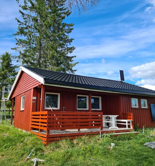 Rød enebolig med nytt mørkt metalltak, terrasse og grønne omgivelser under blå himmel.