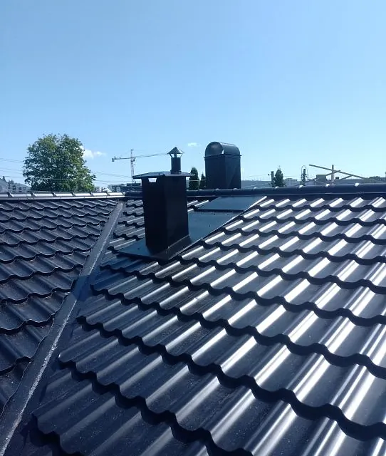 Mørkt metalltak med pipehatter og taksteinprofil, fotografert i sollys mot klar himmel.
