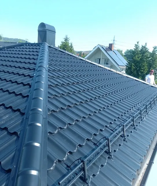 Mørkt metalltak med snøfangere og takhatt, fotografert fra mønet i et boligområde.